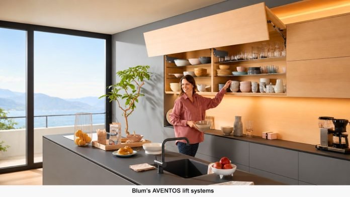 Woman in a pink shirt stands by a modern kitchen island, placing a bowl while open wooden cabinets reveal neatly arranged dishes and glassware with a scenic mountain view through large windows nearby.
